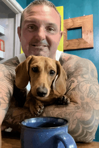 a man holding a dachshund next to a blue cup of coffee