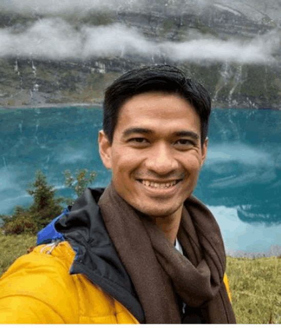 a man in a yellow jacket and scarf smiles in front of a blue lake