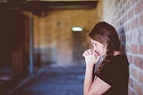 Woman leaning against a brick wall, eyes closed, her mouth resting on clasped hands