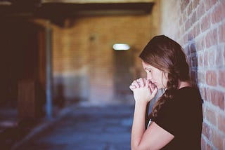 Woman leaning against a brick wall, eyes closed, her mouth resting on clasped hands