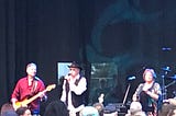 Three performers on stage — left to right, a male bass player (John Billings), a male singer (Micky Dolenz) and a female singer (Coco Dolenz)