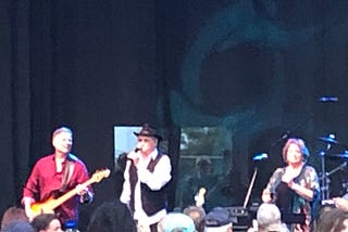 Three performers on stage — left to right, a male bass player (John Billings), a male singer (Micky Dolenz) and a female singer (Coco Dolenz)