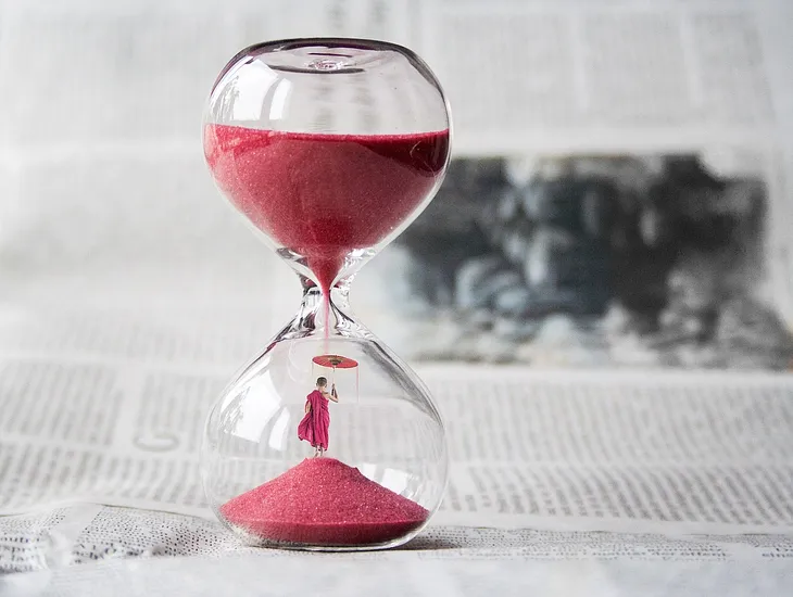 The picture shows an hour glass filled with red sand. A tiny man is at the bottom of the glass holding an umbrella over his head.