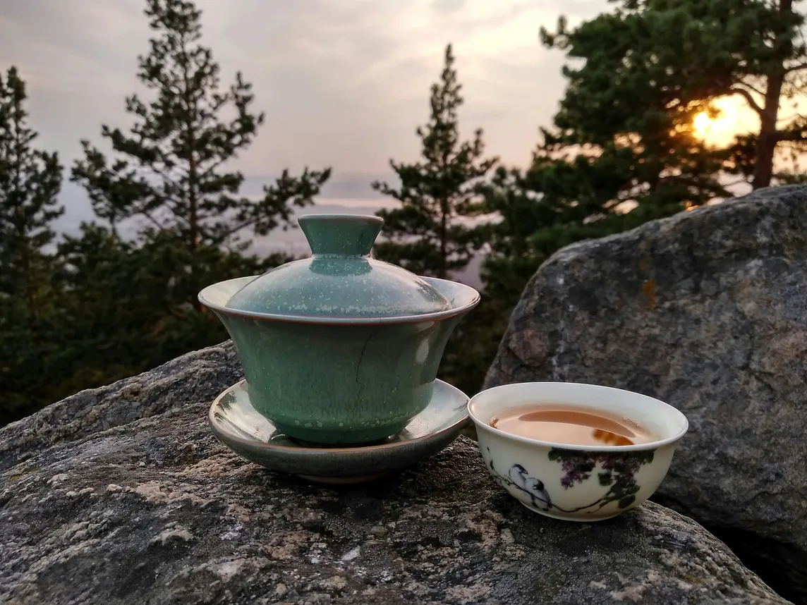 Two bowls of tea, placed on a rock, one open, one covered, with trees and the sea as a backdrop