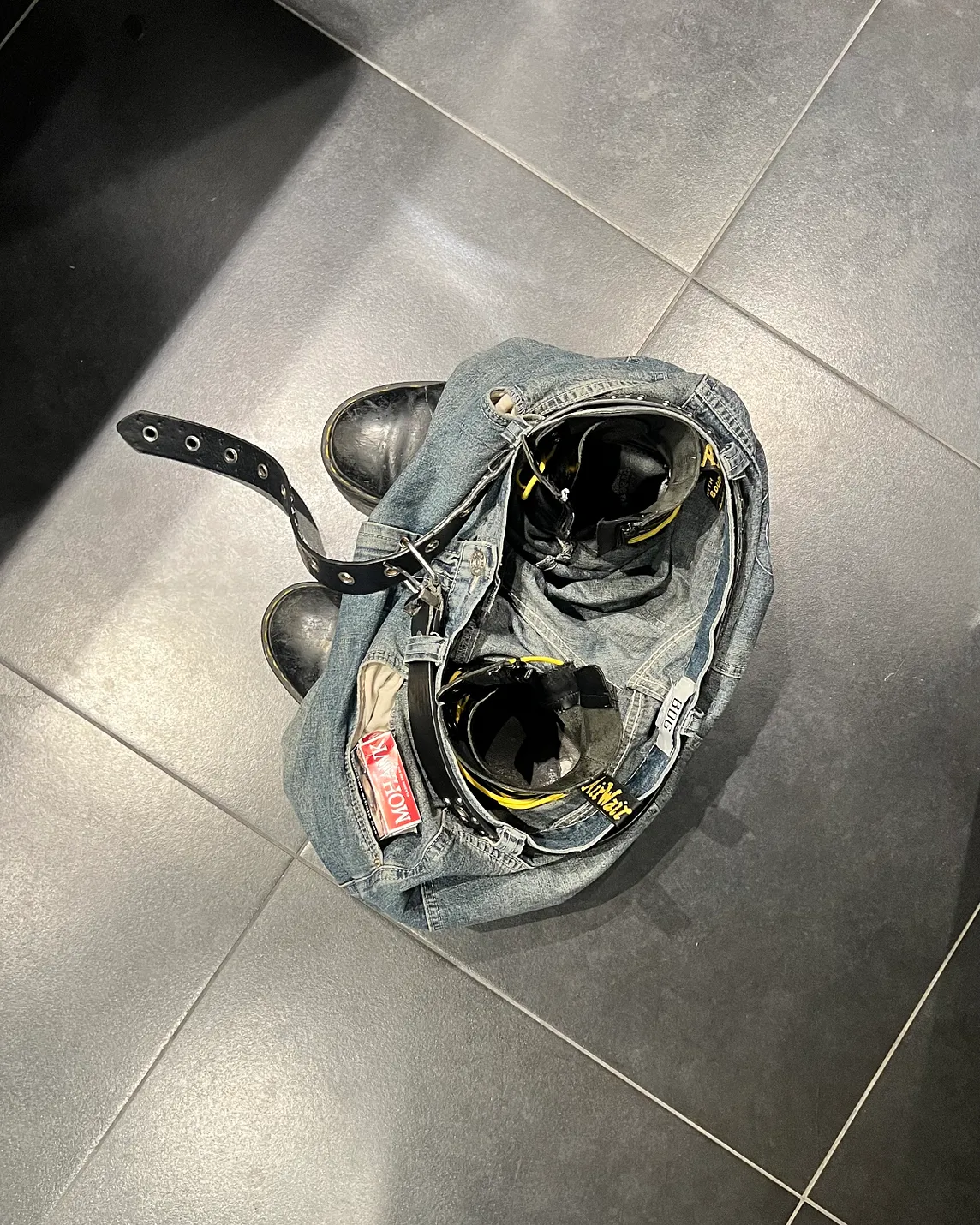 A pair of denim jorts and Doc Martens boots on a tiled floor. The belt is undone and a packet of cigarettes are found in the left pocket.