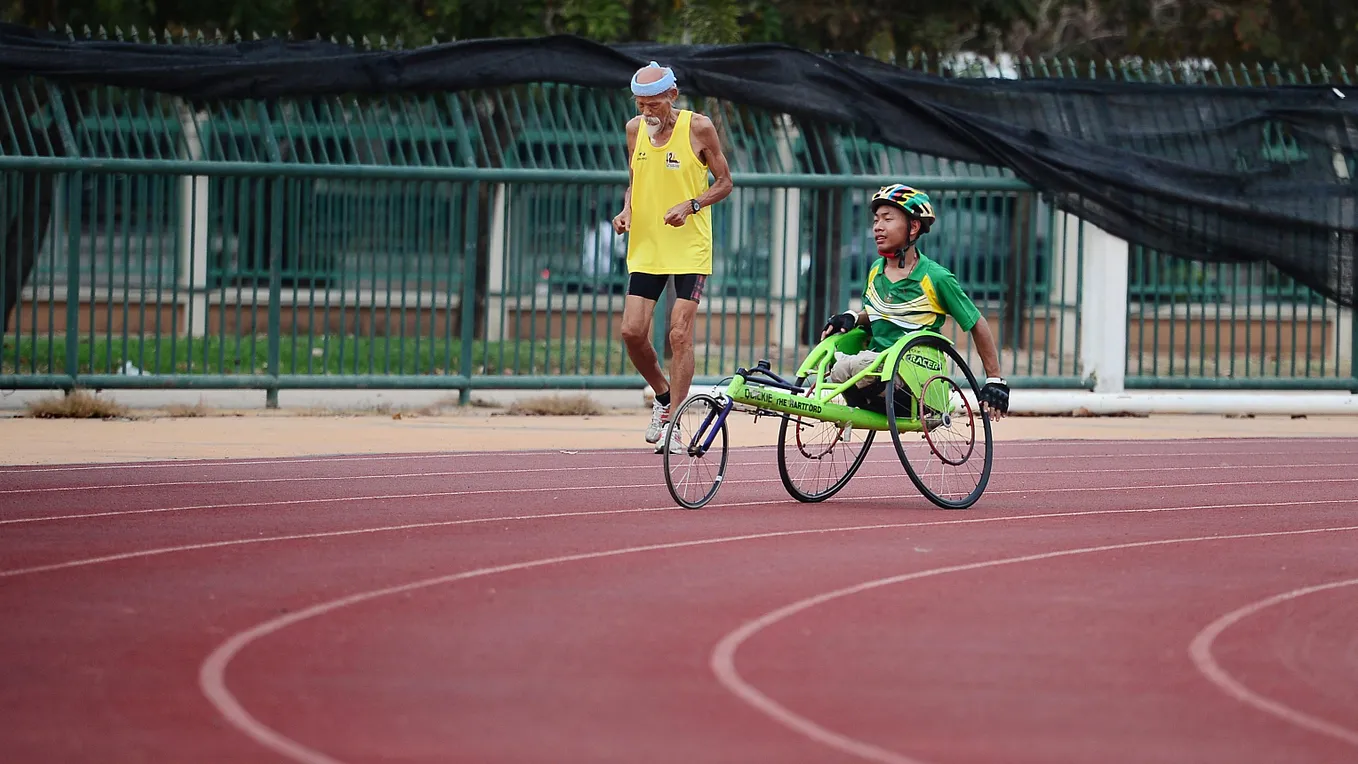 A person on a green racing wheelchair and an abled elderly man running on a track