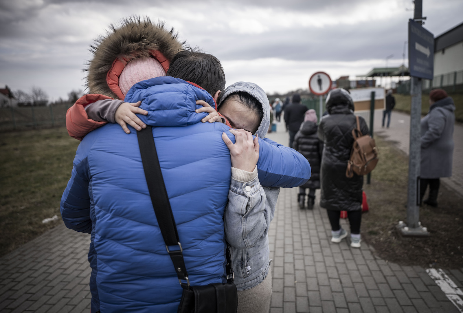 Three people embrace emotionally on a sidewalk, other people can be seen in the background.