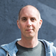 A middle-aged man wearing a hoodie in front of a wall.