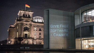 German parliamentary buildings. The Bundestag on the background and in the foreground, another German parliament building with the sentence Give Free Software a chance projected on that builiding walls 