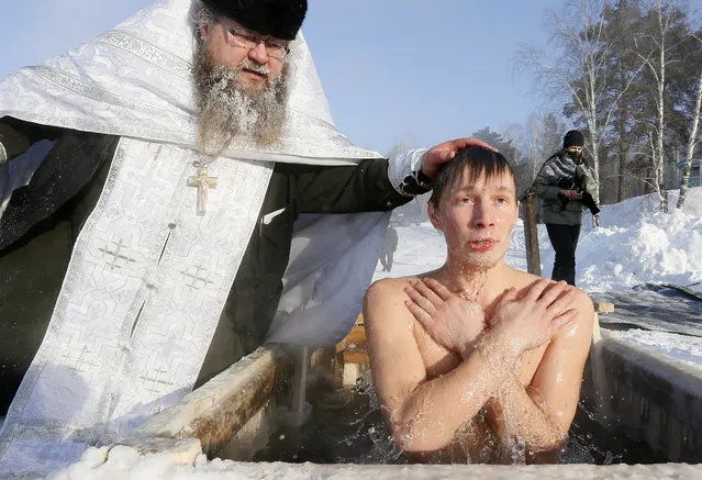 Priest Sergei Ryzhov conducts a ceremony as a man takes a dip in the freezing waters of Lake Buzim during Orthodox Epiphany celebrations near the village of Sukhobuzimskoye north of Krasnoyarsk, Russia on January 19, 2018. (Photo by Ilya Naymushin/Reuters)