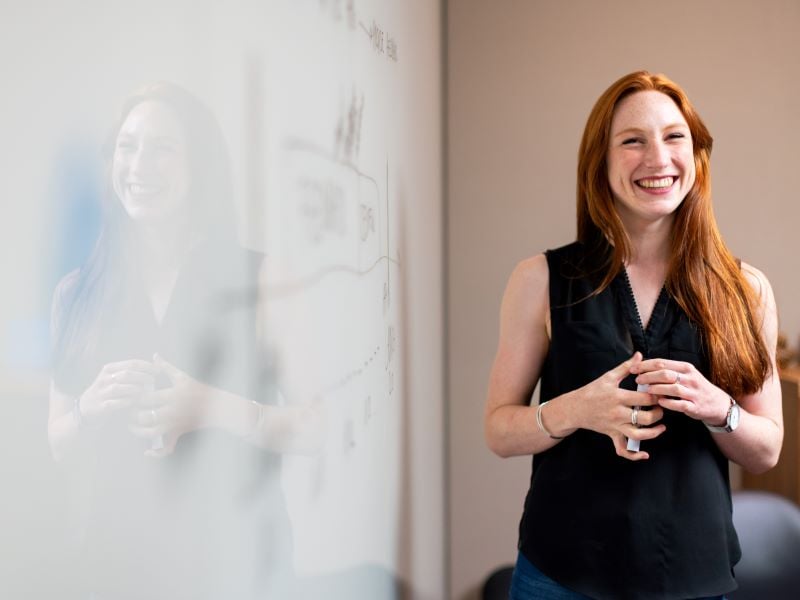 A white female teacher stands at the front of a classroom in front of a whiteboard and holding a whiteboard pen, smiling