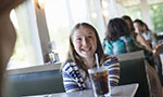 Foto horizontal. Uma menina branca e loira sorrindo e sentada em um assento de restaurante. Na frente um copo de refrigerante com canudo em cima de uma mesa. No fundo outras pessoas sentadas. O cabelo dela é liso e vai até a altura do peito. Usa uma blusa listrada. Do lado dela janelas e muita claridade.