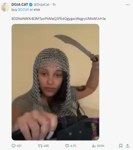 Tweet by Doja Cat: "buy $DOJA or else" followed by a Solana address. There's a photo of her brandishing a toy scimitar and she's wearing a chainmail hood.