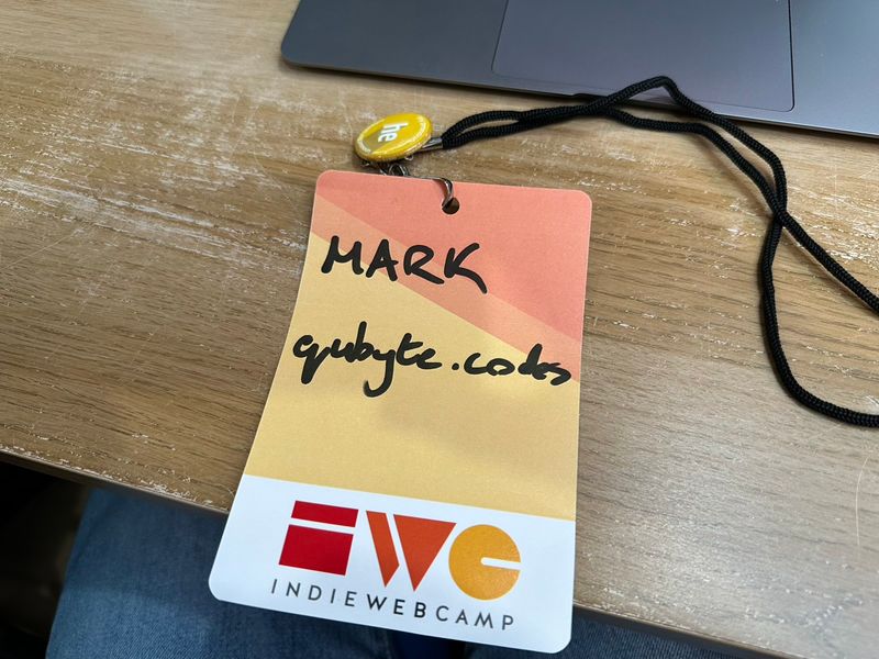 A photograph of my lanyard. It has the IndieWebCamp logo at the bottom, and diagonal yellow, orange, and red bands above. My name and web address are written on in thick black pen.