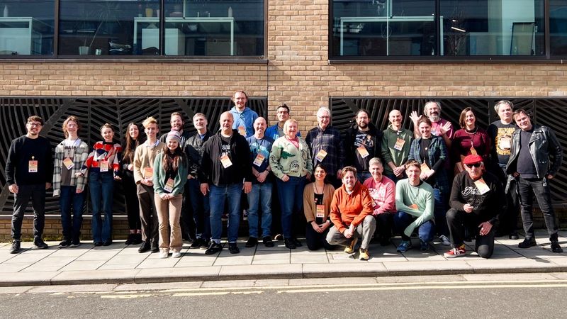 The group photo for Saturday. Twenty six people stood in front of a building. The sun is shining and folk are in shirts, hoodies, and a few jackets.