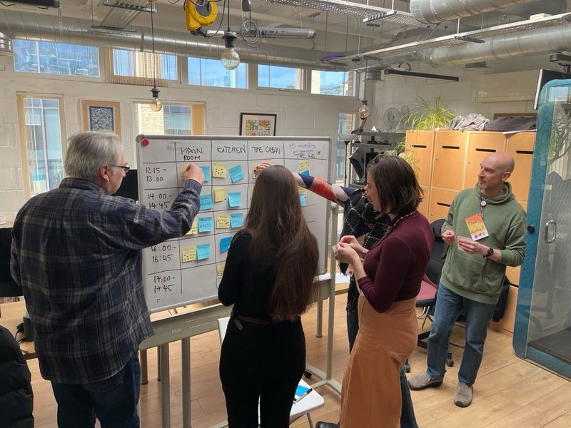Jeremy Keith, Ana Rodrigues, Ros, and Francesco Figari around a whiteboard. The board is split into columns for spaces, rows for times, and each cell has a yellow sticky note for a topic and a blue sticky note with the champion name on.