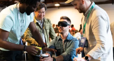 A main holding a coffee cup wearing a blindfold being assisted by three people wearing UX London lanyards