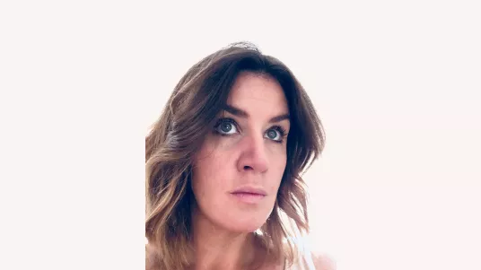 Headshot of a woman with brown wavy hair. She is looking up. On a off-white background.