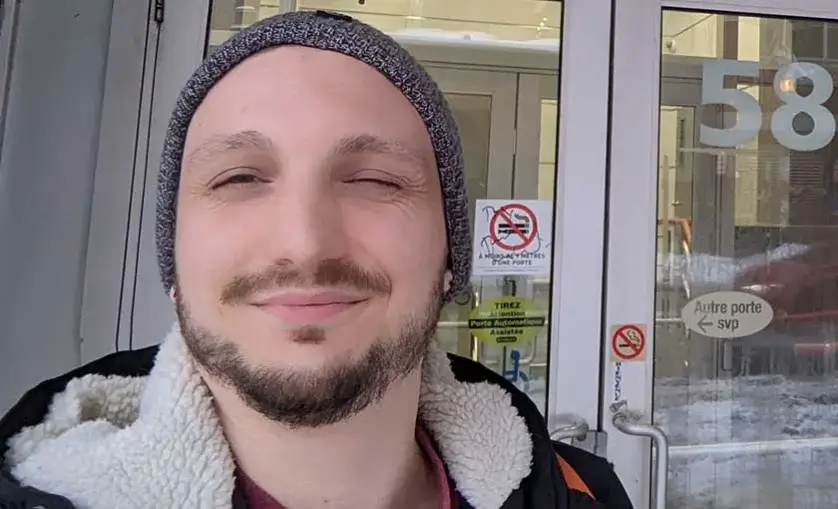 A Person is winking to camera, standing in front of glass doors. They are hearing a hoodie and a beanie.