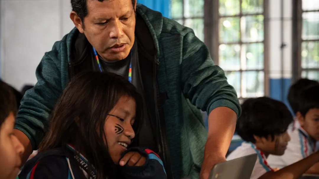 A teacher wearing a green sweatshirt stands over a child at their laptop computer and points to the screen.