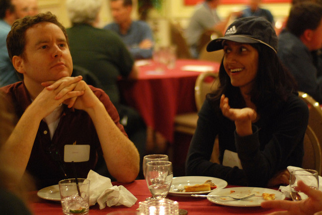 A woman (on the right) and a man (on the left) sitting at a table in a banuet hall. They are both wearing black shirts. The woman is speaking, with a smile and a hand gesturing forward while the man is looking off into the distance, a thoughtful look on his face.