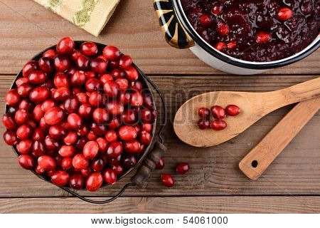 Overhead of a bucket of cranberries and a pot full of whole cranberry sauce on a rustic wooden table. Cranberry sauce is a traditional Thanksgiving side dish. Horizontal format.