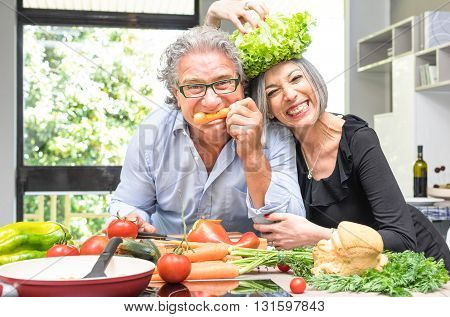 Senior couple having fun in kitchen with healthy food - Retired people cooking meal at home with man and woman preparing lunch with bio vegetables - Happy elderly concept with mature funny pensioner