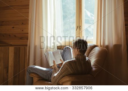 Young man in warm sweater reading book while relaxing on armchair by the window inside cozy log cabin