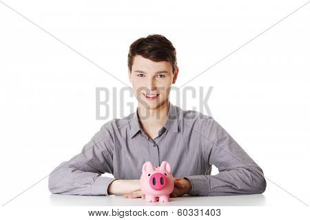 Young man sitting behind the desk with piggy bank on white