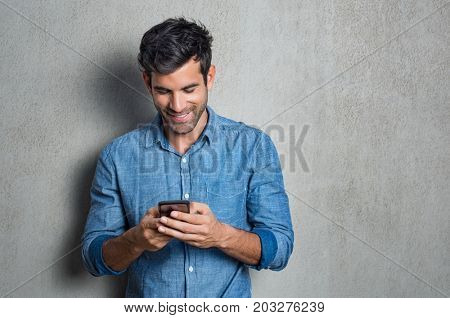 Young man texting message on smart phone isolated on grey background. Smiling latin man holding smartphone and looking at it. Happy hispanic man writing a message on the  phone.