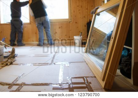 Workers in the background installing new three pane wooden windows in an old wooden house with a new window in the foreground. Home renovation sustainable living energy efficiency concept.