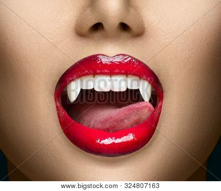 Sexy Vampire Woman's red bloody lips close-up. Vampire girl licking fangs with tongue. Fashion Glamour Halloween art design. Close up of female vampire mouth, teeth. Vampire woman teeth closeup