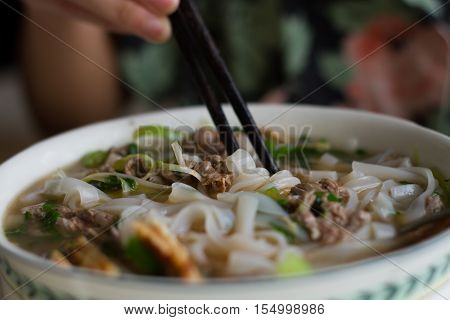 Vietnamese beef noodle soup Pho Bo with garnish of leaves of cilantro, Asian basil and bean sprouts on kitchen table. Young woman is cooking in background. Traditional healthy food.