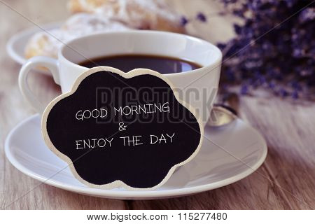 the text good morning and enjoy the day written in a thought bubble-shaped blackboard placed in a cup of coffee, with some pastries in the background in a set table for breakfast