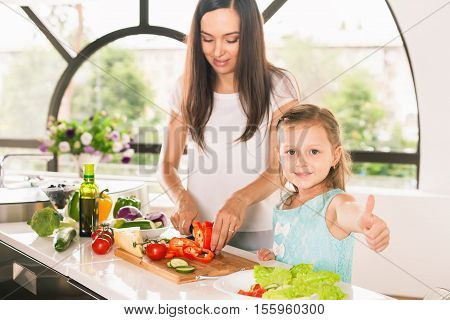 Cute little girl cooking with her mother and shows thumbs up. Healthy food, cooking healthy salad with vegetables ingredients. Mom and daughter cooking together. Recipe food for baby or child