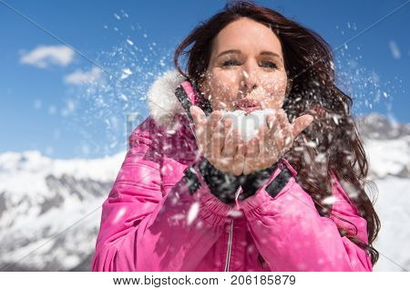 Beautiful young woman blowing snow from her hands in winter. Closeup face of brunette woman playing with snow at ski resort. Happy girl holding snow in hand and blowing it on snowy mountain.