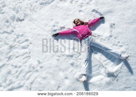 High angle view of happy woman lying on snow and moving her arms and legs up and down creating a snow angel figure. Smiling woman lying on snow in winter holiday with copy space.