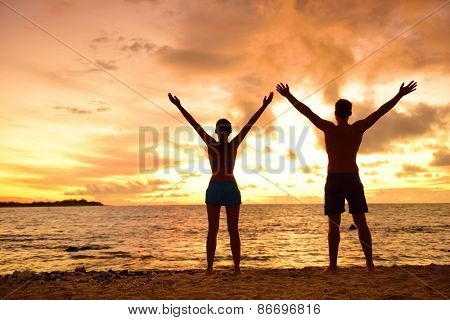 Freedom people living a free, happy, carefree life at beach. Silhouettes of a couple at sunset arms raised up showing happiness and a healthy lifestyle against a colorful sky of clouds background.