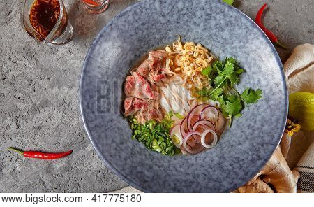 Vietnamese Cuisine - Beef Pho Noodle Soup or Pho Bo Soup. Served with Fresh Greens in Blue Bowl on Textured Dark Gray Table. Pho Bo Soup with Spicy Sauces. Top View