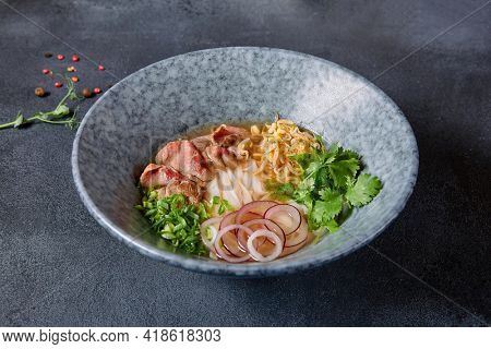 Vietnamese Cuisine - Beef Pho Noodle Soup or Pho Bo Soup. Served with Fresh Greens in Blue Bowl on Textured Dark Gray Table. Pho Bo Soup with Spicy Sauces