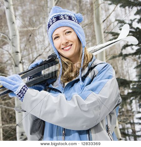 Mid adult Caucasian female skier wearing blue ski clothing carrying skis on shoulder.