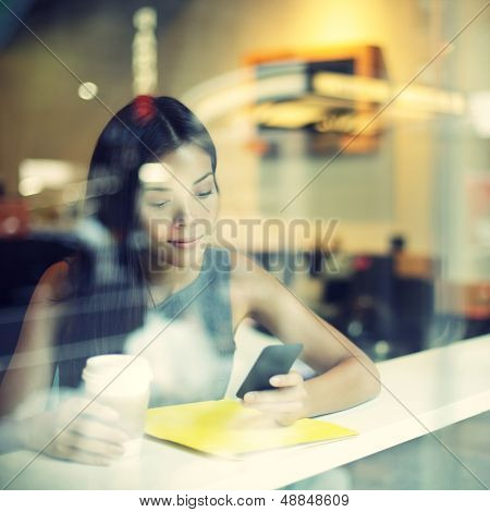 Cafe city lifestyle woman on phone drinking coffee texting text message on smartphone app sitting indoor in trendy urban cafe. Cool young modern mixed race Asian Caucasian female model in her 20s.