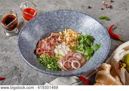 Vietnamese Cuisine - Beef Pho Noodle Soup or Pho Bo Soup. Served with Fresh Greens in Blue Bowl on Textured Dark Gray Table. Pho Bo Soup with Spicy Sauces