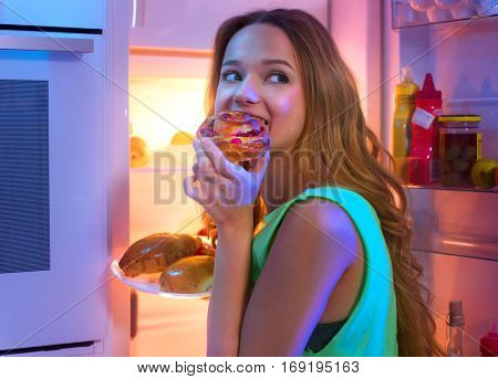 Portrait Of Beautiful Teenage girl Taking Food From Refrigerator at night. Night eating, overeat concept. Diet, dieting. Beauty young woman eating pastries in fridge at night kitchen.