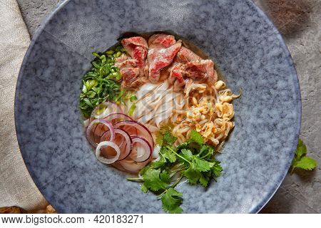 Vietnamese Cuisine - Beef Pho Noodle Soup or Pho Bo Soup. Served with Fresh Greens in Blue Bowl on Textured Dark Gray Table. Pho Bo Soup with Spicy Sauces. Top View