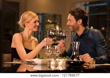 Romantic young couple at restaurant raising a toast. Beautiful couple with glasses of red wine in restaurant. Couple toasting wine glasses during a romantic dinner in a gourmet restaurant.
