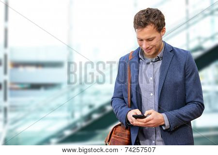 Young urban professional man using smart phone in office building indoors. Businessman holding mobile smartphone using app texting sms message wearing suit jacket and bag.