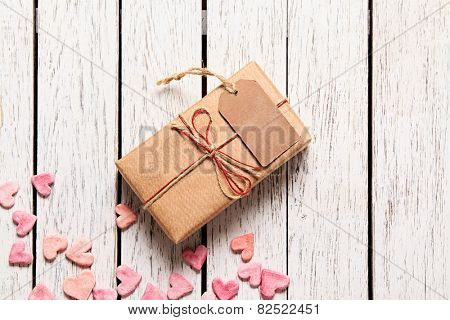 Gift box with blank gift tag and heap of small hearts on white wooden background.