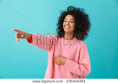 Image of american curly woman in pink shirt looking aside and pointing index finger on copyspace with smile isolated over blue background