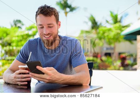 Smartphone man sms texting drinking cold coffee drink at outdoor cafe on terrace in summer. Happy casual urban young adult holding phone reading or using app to connect with friends on social media